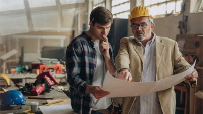 Young carpenter and mature quality control inspector reading blueprints in a factory.