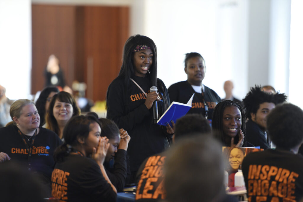 A college student, holding a notebook, speaks into a microphone. She is surrounded by a cheering group of young people wearing matching black and orang