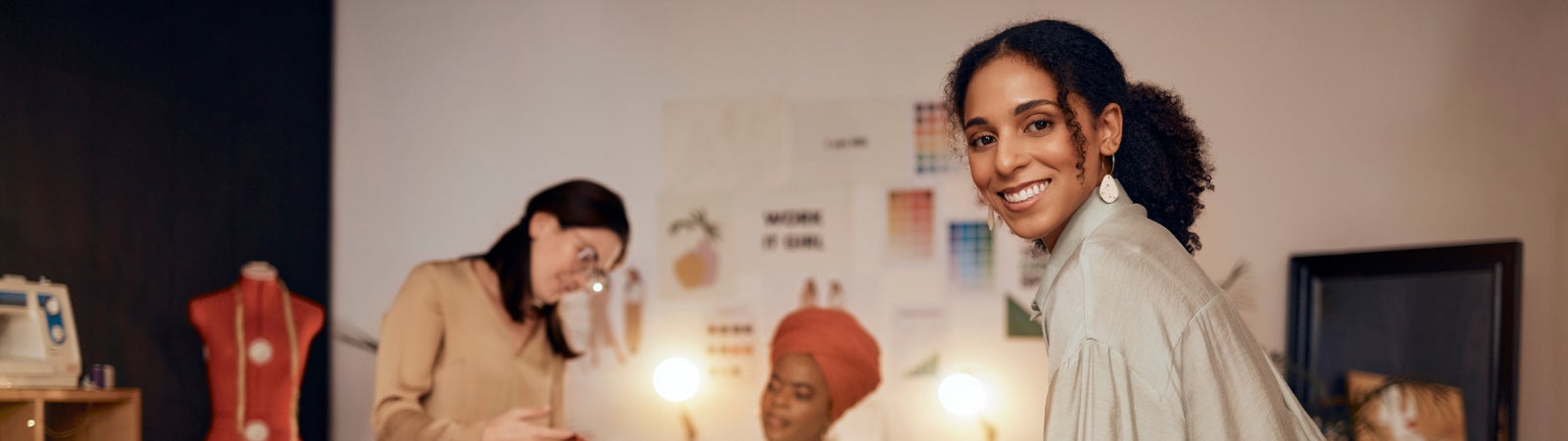 A photo of a fashion studio, including a Black small business owner smiling toward the camera with color swatches laid out on the desk in front of her, while her coworkers discuss a garment in the background.