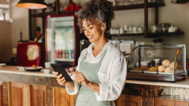 Young female worker holding a digital tablet, smiling while standing with a cafe shop in the background.