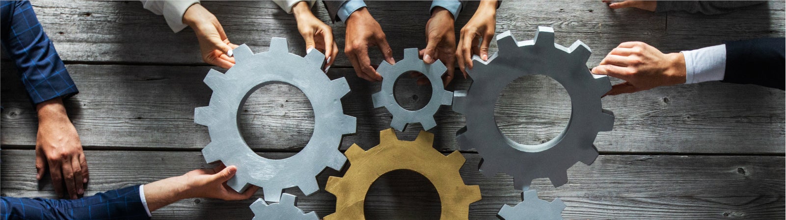 Photo of gears on a table, held together by the hands of people seated around it.