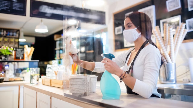 Photo of a young woman with a face mask working in a cafe, wiping down a plastic barrier.