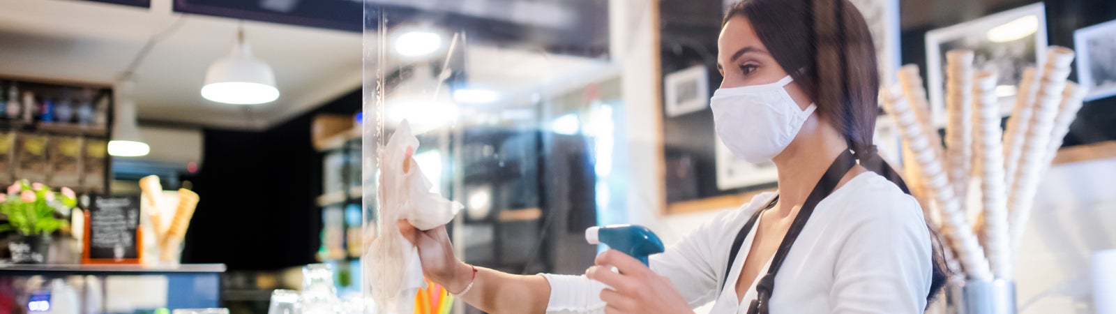 Photo of a young woman with a face mask working in a cafe, wiping down a plastic barrier.