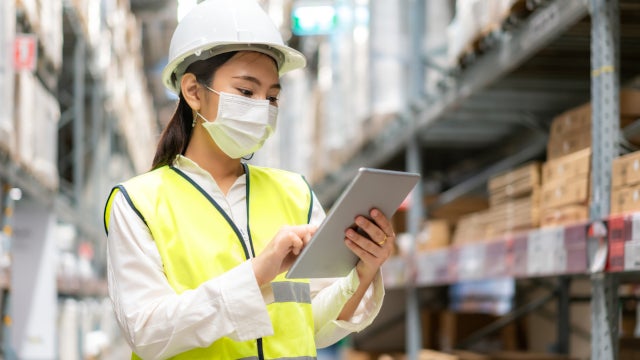 Photo of an Asian woman working in a warehouse. She is using a tablet and wearing a hard hat, face mask, and reflector vest.