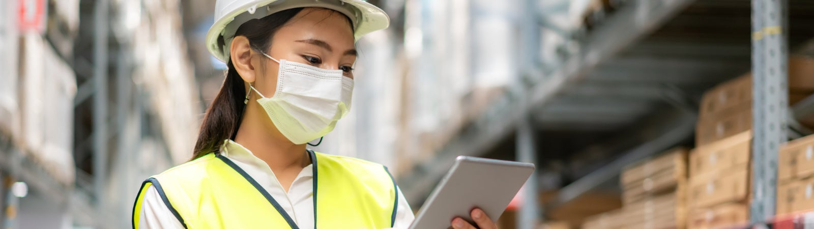 Photo of an Asian woman working in a warehouse. She is using a tablet and wearing a hard hat, face mask, and reflector vest.