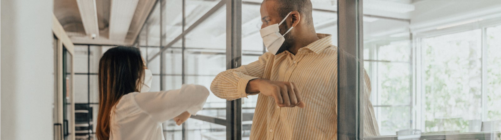Photo of a Black man and Asian woman wearing face masks and bumping elbows in an office.