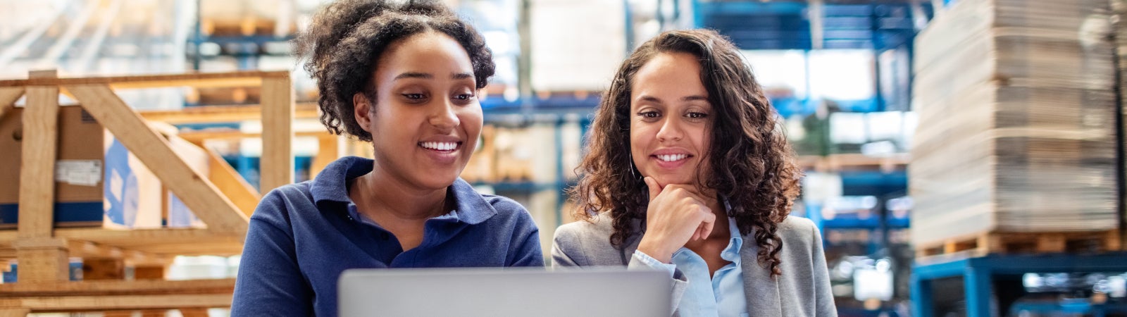 Photo of a young woman worker and woman supervisor in a warehouse. They are smiling while using a laptop.