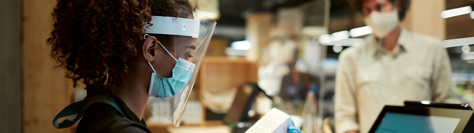 Photo of a young woman of color working at the checkout lane in a grocery store, wearing personal protective equipment, including a face mask, face shield, and gloves