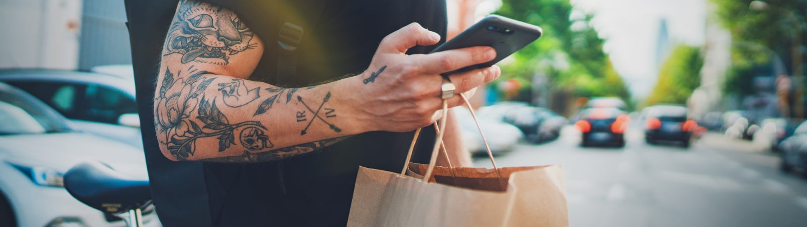Photo of a man using an app on his smartphone to complete a delivery. He is riding a bicycle while carrying a backpack and a paper shopping bag.