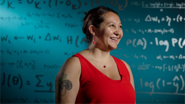 A woman in a red sleeveless top stands in from of a wall of mathematical equations. Photo credit: Jordan Stead / Amazon.