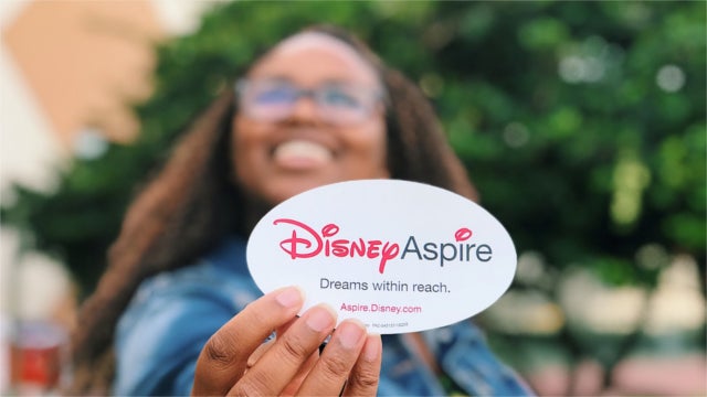 Disney employee holds up a sticker that says "Disney Aspire. Dreams within reach. Aspire.Disney.com"