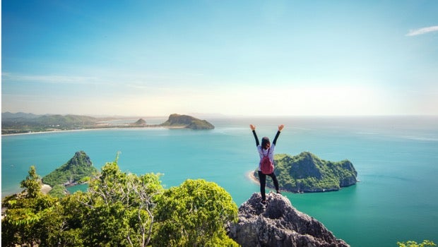 A woman standing on top of a mountain overlooking water with her arms raised.