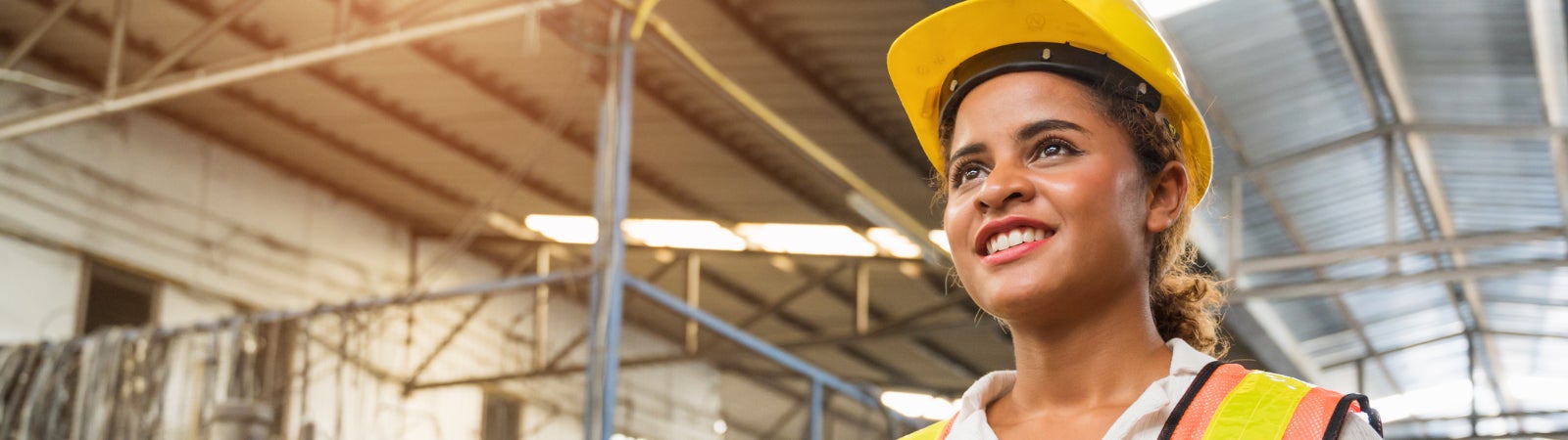 Photo of a young woman factory worker wearing a hard hat and reflector vest. She is smiling and holding an electronic tablet.