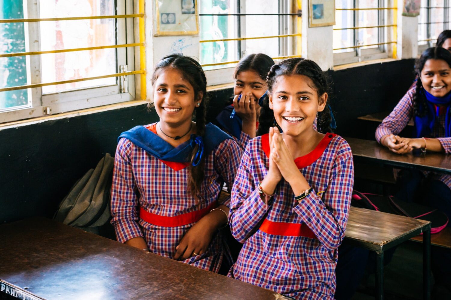 A group of young students sit at wooden desks smiling at the camera.